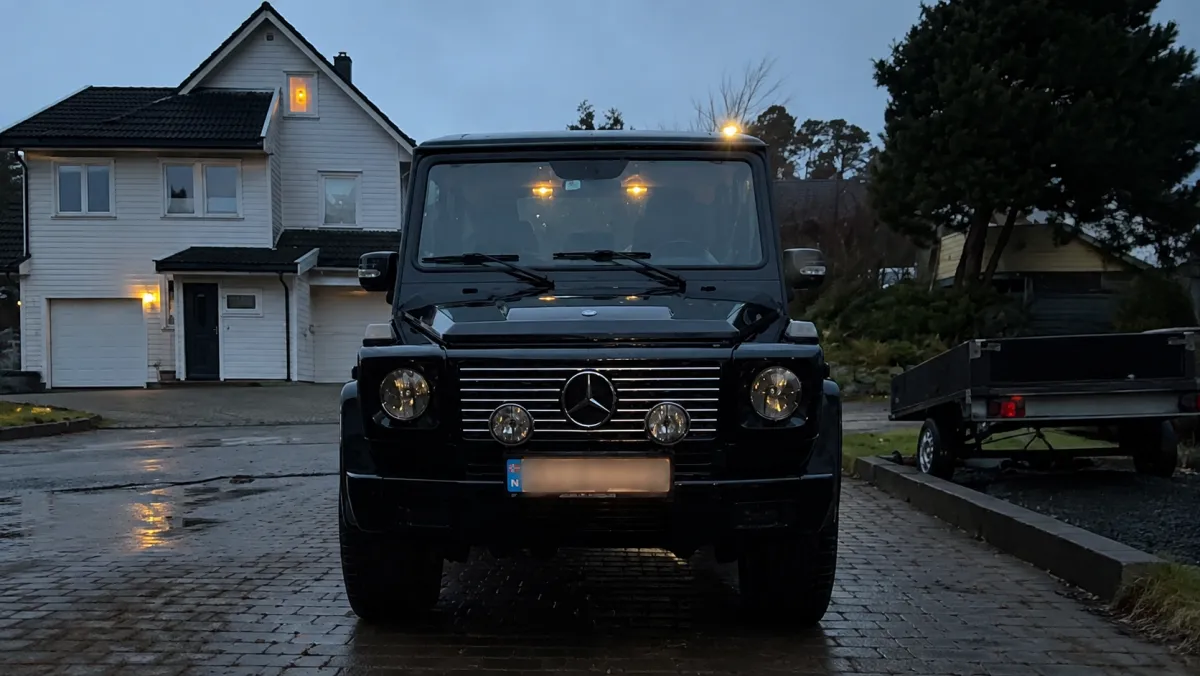 Nyvasket svart Mercedes G-klasse på våt brostein foran et bolighus i skumringen